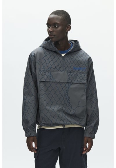 Jeune homme portant une veste à capuche grise avec un motif à maillons bleus et un pantalon cargo noir, regardant vers sa gauche.