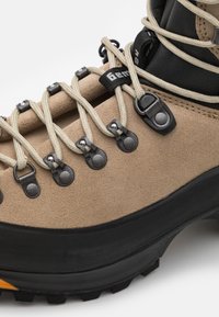 Botă de drumeție din suede bej cu talpă din cauciuc negru, dotată cu gromete metalice și sireturi bej, concepută pentru durabilitate și aderență.