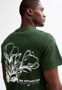 Grön bomullströja med en vit blommig grafik på baksidan, med texten "GREAT OUTDOOR CLUB" och en texturerad tygfinish.