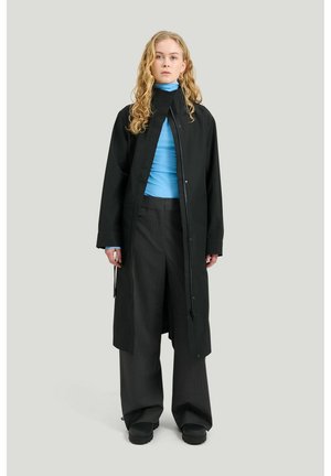 Vaaleahiuksinen nainen, joka pukeutunut pitkään mustaan takkiin, vaaleansiniseen poolopaitaan, leveisiin mustiin housuihin ja mustiin kenkiin, seisoo yksinkertaista vaaleaa taustaa vasten.