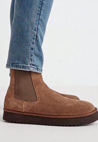 Botines de ante marrón con paneles laterales elásticos, punta redonda y suela de goma texturizada. Combinados con jeans de color azul claro.