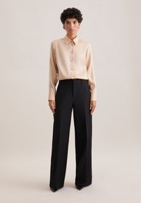 Camicia button-up beige in satin con maniche lunghe, abbinata a pantaloni neri a vita alta e a gamba larga. Collo slim fit e design minimalista.