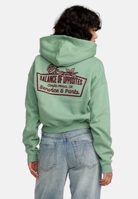 Sweat à capuche vert avec un grand design imprimé au dos représentant des roses et du texte. Manches et taille côtelées, associé à un jean en denim clair.