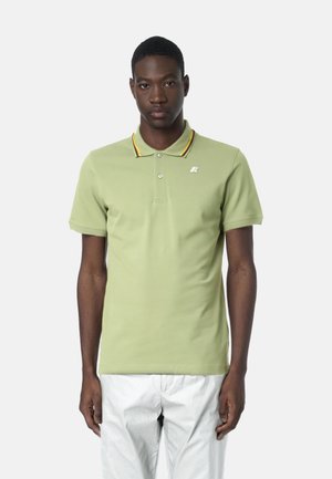 Uomo che indossa una polo verde chiaro con righe multicolori sul colletto e pantaloni bianchi, in piedi di fronte a uno sfondo semplice.