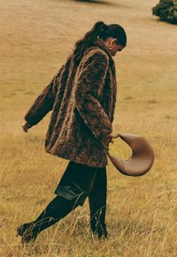 Aizstātā kažokādas brūns mētelis ar garām piedurknēm, uzvilkts virs melnas ādas svārku un līdz ceļiem melnām zābakiem. Nēsā izliekta brūna rokas soma.