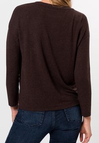 Haut marron à manches longues, coupe décontractée avec ourlet arrondi, fabriqué dans un tissu doux, présentant une texture subtile. Porté avec un jean en denim.