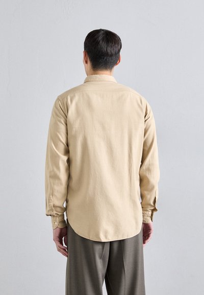 Beige långärmad skjorta med knäppningar vid ärmsluten, lätt tyg, rak passform och rundad nederkant. Slät textur med minimal dekoration.