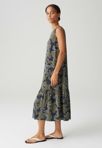 Sleeveless, tiered dress in olive green with navy abstract pattern and white outlines. Soft fabric with a relaxed fit, paired with flat sandals.