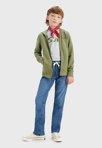 Felpa verde con zip, t-shirt grigia con grafica, jeans blu e sneakers bianche con accenti blu navy. Bandana rossa attorno al collo.