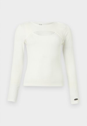 Λευκό γυναικείο τοπ με μακριά μανίκια, στρογγυλεμένη κοπή στο μπροστινό μέρος και μικρό μεταλλικό λογότυπο στο μανσέτα του αριστερού μανικιού.