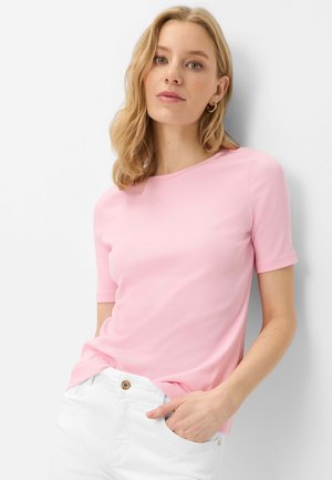 Blond kvinde iført en lyserød kortærmet top og hvide bukser, stående med den ene hånd i lommen mod en hvid baggrund.