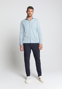 Lichtblauwe overhemd met lange mouwen en een gladde textuur, gecombineerd met donker marineblauwe slim-fit broeken en casual witte sneakers.