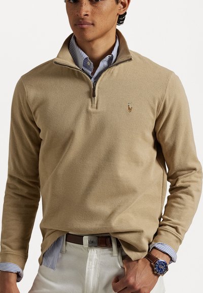 Beige half-zip pullover made of textured fabric, featuring a small embroidered logo on the chest and layered with a blue and white striped shirt.