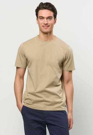 Nuori mies, jolla on tummat hiukset, beige lyhythihainen t-paita ja tummansiniset housut, seisoo yhdellä kädellä taskussa ja hymyilee kameralle.