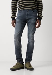 Slim-fit farkut tummansinisinä, kevyellä kulutuksella, perinteisellä viiden taskun suunnittelulla, yhdistettyinä oliivinvihreisiin lenkkikenkiin.