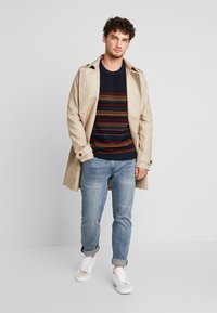 Beige Trenchcoat über einem gemusterten marineblauen Pullover, kombiniert mit hellblauen Jeans und weißen Sneakern. Verfügt über eine taillierte Passform und Knopfdetails.