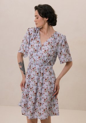 Sukienka w stylu wrap w jasnoniebieskim materiale z kwiatowym wzorem, który zawiera czerwone i zielone akcenty. Krótkie rękawy i zapinana w talii, z luźną spódnicą.