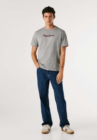 Tricou din bumbac gri cu mâneci scurte, având logo-ul roșu "Pepe Jeans London", asortat cu blugi albaștri și adidași bej.