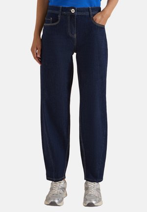 Person iført mørkeblå jeans med lige ben, en blå top og lysegrå sneakers, stående med den ene hånd i lommen.