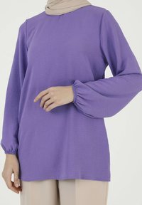 Purple long-sleeve top made of lightweight, textured fabric, featuring elasticized cuffs and a relaxed fit, paired with beige trousers.