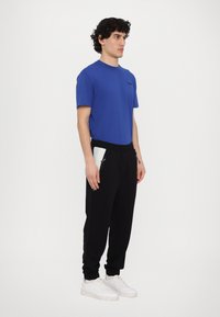 Blå bomulds t-shirt, sorte sweatpants med sidelommer med lynlås, model iført hvide sneakers, stående mod en hvid baggrund.
