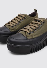 Baskets en toile vert olive avec embouts en caoutchouc noir et semelles épaisses. Présente des lacets beiges et des œillets en métal pour un maintien sécurisé.