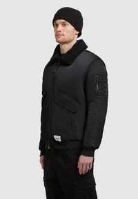 Sort bomberjakke med en faux fur-krave, lynlåslommer, ribbede manchetter og en tekstureret overflade. Model iført en sort strikhue.
