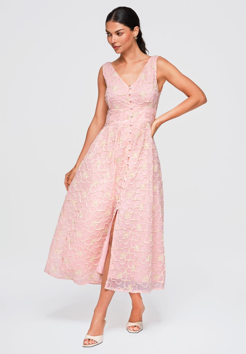 Femme portant une robe midi rose sans manches avec broderies florales et boutons sur le devant, posant avec une main sur la hanche et des talons blancs.