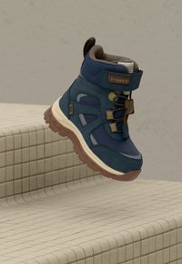 Botte de randonnée pour enfant bleu et marron avec fermeture Velcro et lacets, présentée flottant au-dessus de marches carrelées beige sur fond gris uni.