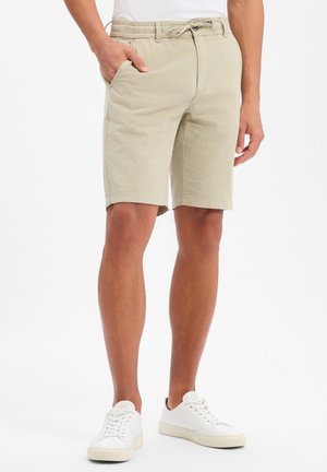 Homme portant un short beige mi-long, taille élastique avec cordon, baskets blanches, debout avec une main dans la poche.