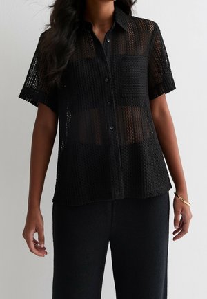 Musta lyhythihainen paita, jossa on teksturoidtu, avoin neulontakuvio ja etutasku. Napitettava, rento istuvuus ja pehmeä kangas.