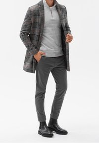 Grå ternet frakke med orange detaljer over en lys grå zip-up sweater, parret med grå bukser og sorte ankelstøvler.