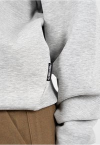 Grijze sweatshirt van zacht materiaal, met een zwart label waarop de tekst "PROHIBITED" staat. Close-up van de details van de mouw en de kraag tegen een achtergrond van bruine broek.