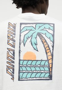 Άτομο που φοράει λευκό πουκάμισο με πολύχρωμο γραφικό ενός φοίνικα, κυμάτων του ωκεανού και ηλιοβασιλέματος, με το κείμενο "HAWAII" στην πλάτη.