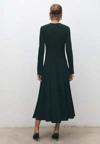 Vestido de manga larga en verde oscuro con falda acampanada, con cremallera en la parte trasera y textura suave. Llevado con tacones altos negros.