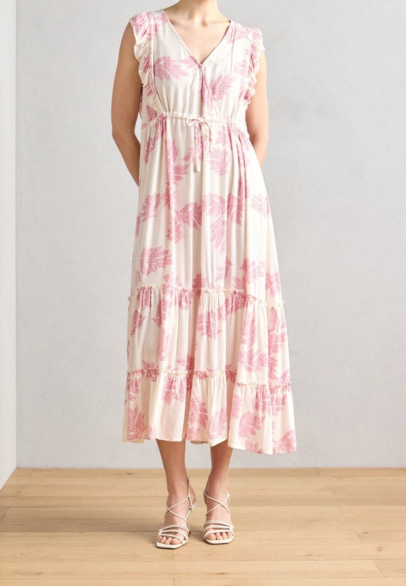 Femme portant une robe midi crème sans manches avec des motifs de feuilles roses, des détails à volants et une taille nouée, associée à des sandales blanches à brides.