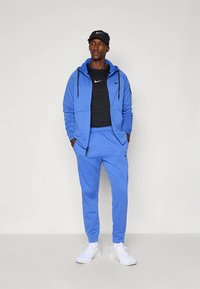 Plavi trenirka od mekanog materijala, s kapuljačom na patentni zatvarač i joggericama s elastičnim strukom, uparena s crnom majicom i bijelim tenisicama.