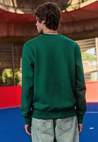 Donkergroen sweatshirt met ronde hals en geribbelde manchetten, gecombineerd met lichtblauwe denim jeans, gezien vanaf de achterkant.