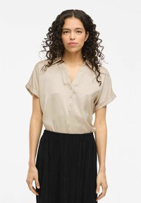 Camicia di satin beige con scollatura a V, maniche corte arrotolate e dettagli con bottoni, abbinata a una gonna plissettata nera. Texture liscia, design elegante.