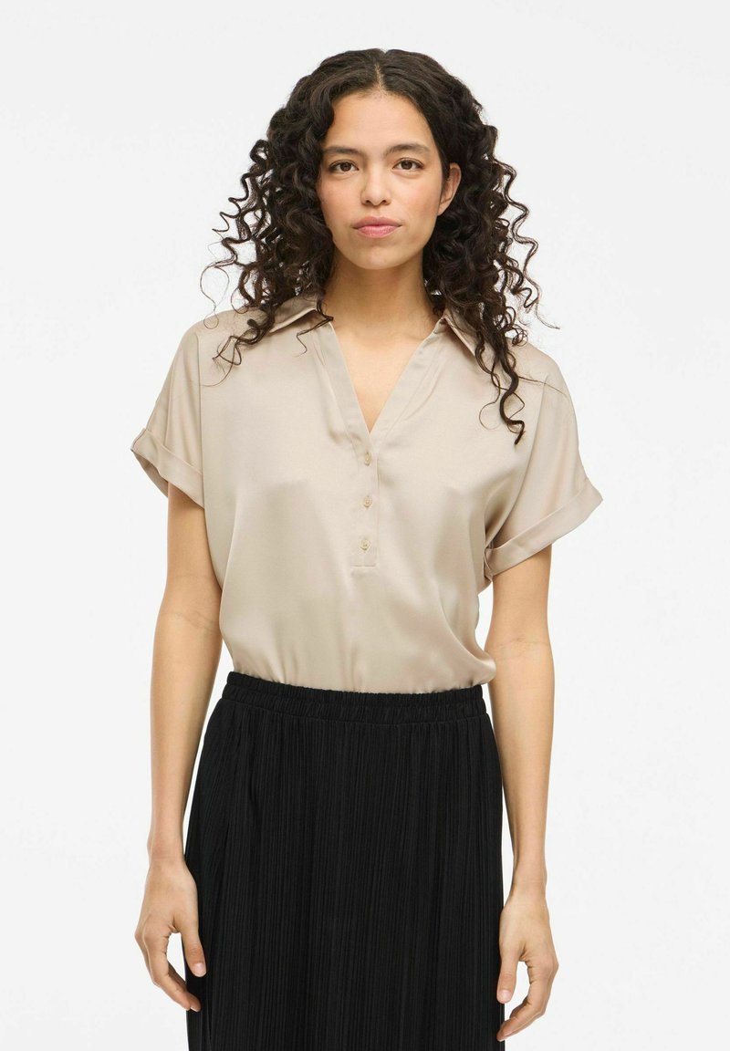 Camicia di satin beige con scollatura a V, maniche corte arrotolate e dettagli con bottoni, abbinata a una gonna plissettata nera. Texture liscia, design elegante.