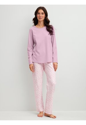 Woman with long dark hair wearing a light purple long-sleeve top and white pants with a pink leaf pattern standing barefoot.
