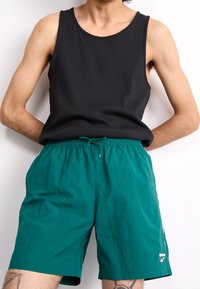 Černý žebrovaný top na ramínka s širokými popruhy, kombinovaný s tyrkysovými šortkami na šňůrku. Šortky mají boční kapsy a malé logo Reebok na lemu.