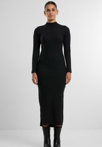Abito lungo nero a maniche lunghe con scollatura alta, silhouette aderente e lunghezza fino alla caviglia. Presenta una texture a costine e un design minimale.
