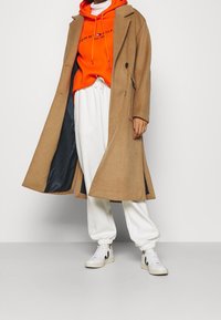 Bruine wollen jas met dubbelbreasted ontwerp, oranje capuchontrui, witte joggingsbroek en witte sneakers met zwarte accenten. Gladde textuur, ontspannen pasvorm.