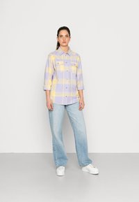 Langermet skjorte i lys lilla og gul rutemønster med to brystlommer, knappelukking, sammen med vide, falmede blå jeans og hvite joggesko.