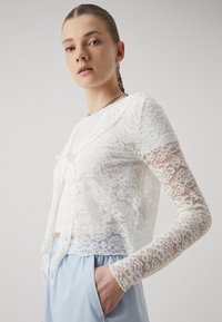 White lace cropped cardigan with long sleeves and floral pattern, tied at the front. Worn over a white top, paired with light blue bottoms.