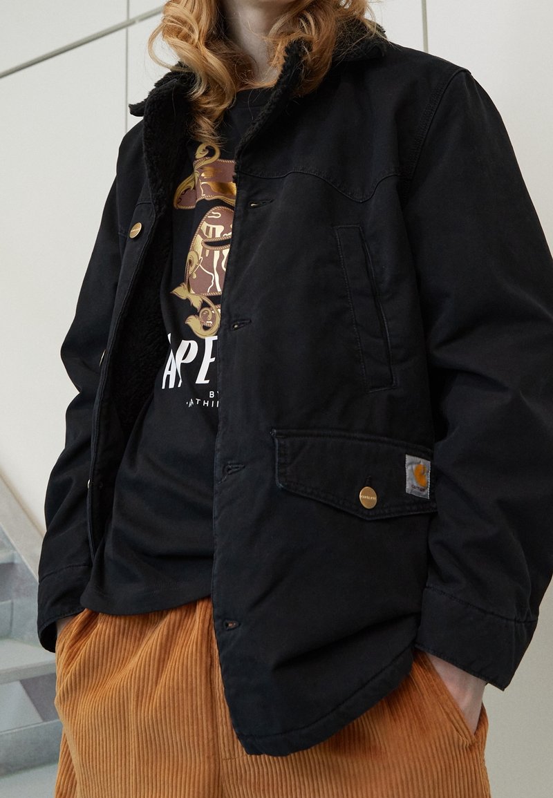 Personne portant une veste noire avec un t-shirt noir graphique et un pantalon en velours côtelé marron, debout à l'intérieur près des escaliers.