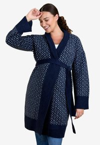 Femeie zâmbitoare cu păr creț poartă un cardigan bleumarin cu curea, cu un model de diamante albe și margini cu nervuri, peste blugi.