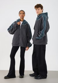 Siv pulover z flisom in turkiznimi detajli ter belimi obrobami, z zadrgo in oversized kapuco, oblečen z črnimi širokimi hlačami in čevlji.