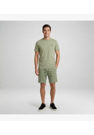 Man staat en lacht, draagt een lichtgroen t-shirt en bijpassende shorts met zwarte sneakers tegen een effen grijze achtergrond.
